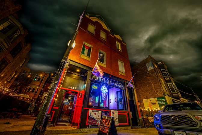 Nighttime urban corner bar in a three-story red brick building with neon open sign, colorful string lights and flags, an open red door and sidewalk chalkboard reading “Paws Welcome,” a parked pickup, and dramatic stormy clouds overhead.