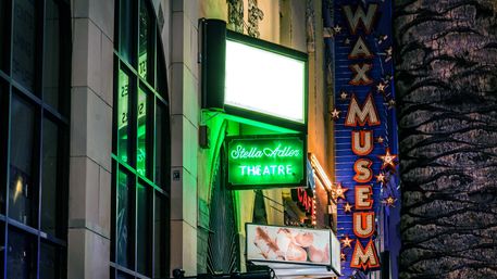 Glowing green neon theatre sign and tall blue star-studded vertical marquee lighting a nighttime city sidewalk, reflected in glass windows beside a textured palm tree trunk.