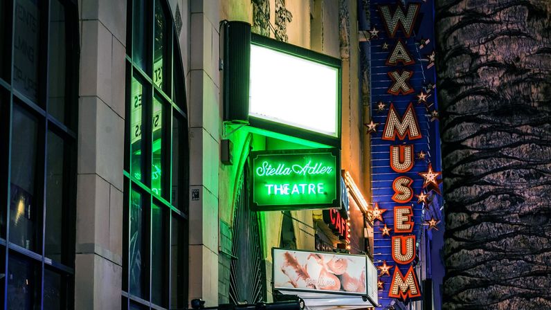 Glowing green neon theatre sign and tall blue star-studded vertical marquee lighting a nighttime city sidewalk, reflected in glass windows beside a textured palm tree trunk.