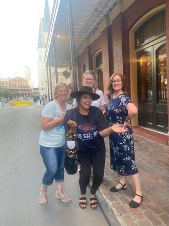 Four women laughing and posing on a lively downtown historic-district sidewalk with brick storefronts and an awning, one wearing a wide-brimmed hat and holding a lantern.