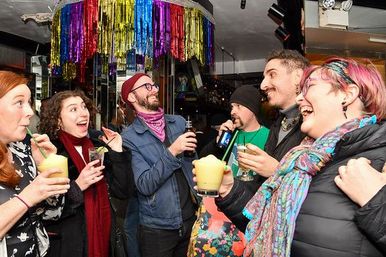 Group of adults laughing and toasting colorful frozen cocktails in a festive indoor bar with rainbow streamers and mirrored walls, lively nightlife scene.