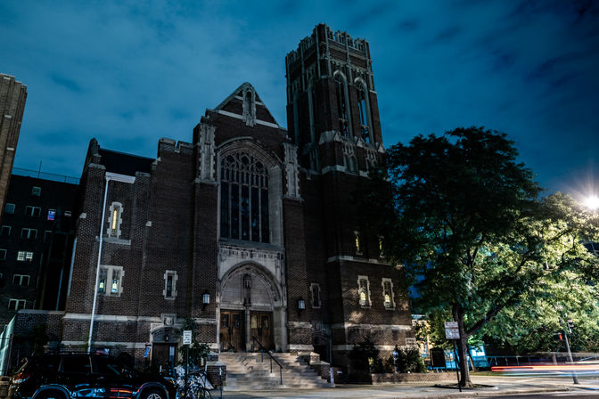 Moody nighttime view of a historic Gothic-style brick church with a tall tower on a downtown street, lit steps, a leafy tree and parked cars under a deep blue sky.