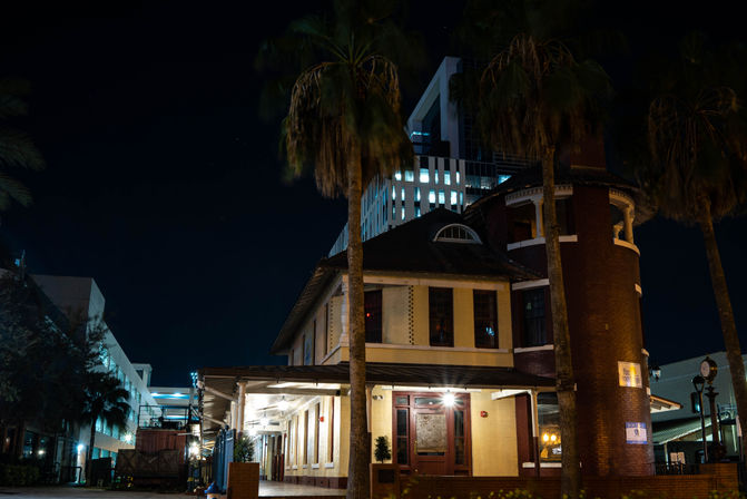 Historic brick-and-stucco building lit at night with palm trees in the foreground and a modern office tower rising behind it in an urban streetscape