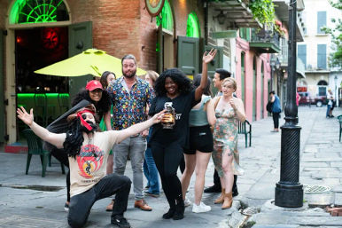 Group of friends cheering and laughing on a lively New Orleans street with colorful buildings, hanging balconies, and a green-lit bar entrance