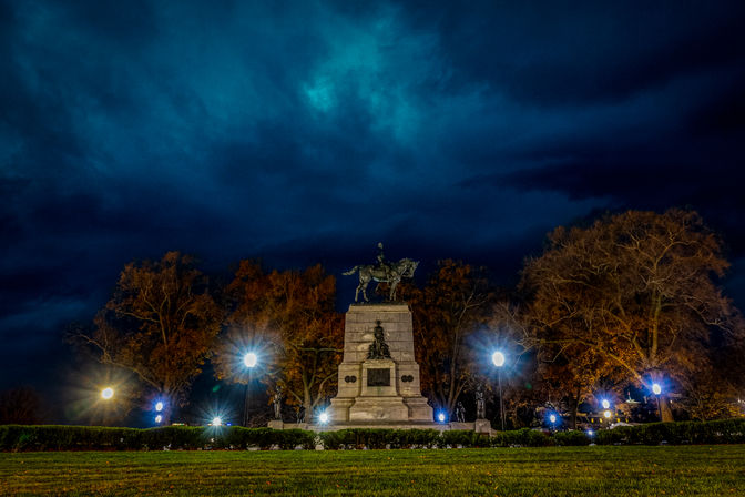 Equestrian statue on a stone monument in a park at night, glowing lampposts and autumn trees under dramatic teal-blue storm clouds