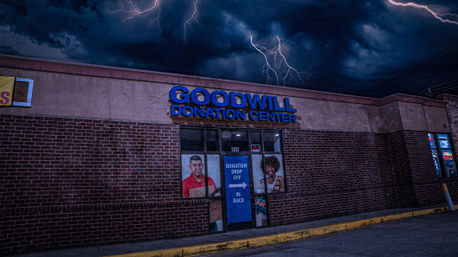 Brick-front charity donation center storefront at night with a blue-lit sign above glass doors and poster figures in the windows, dramatic thunderstorm sky with lightning bolts overhead