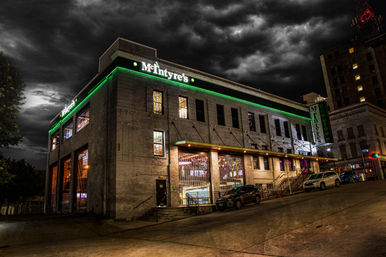 Moody night view of a two-story historic brick bar with green neon trim on a sloped downtown street, glowing interior lights and parked cars beneath dramatic storm clouds.