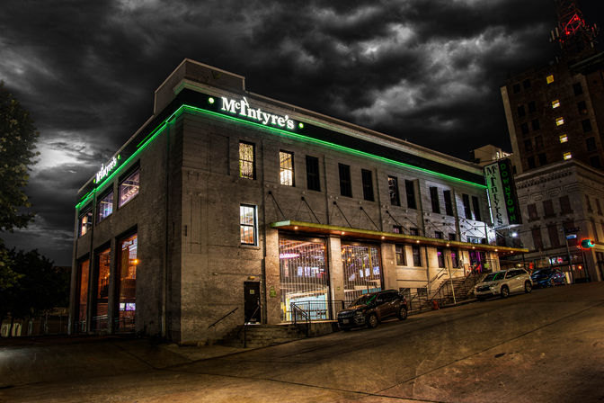 Moody night view of a two-story historic brick bar with green neon trim on a sloped downtown street, glowing interior lights and parked cars beneath dramatic storm clouds.