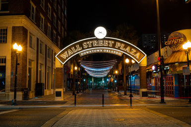 Lit archway entrance to a downtown Orlando pedestrian plaza at night, twinkling string lights over an empty brick-paved street flanked by historic buildings and glowing lampposts.