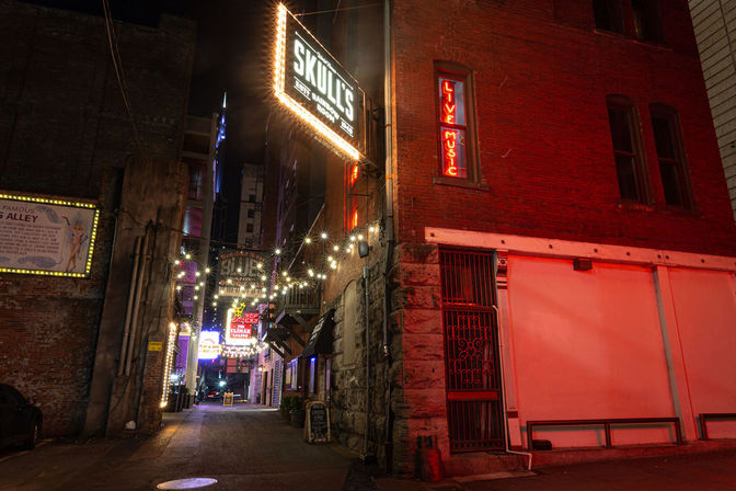 Moody downtown alley at night with brick buildings, colorful neon and marquee signs, overhead string lights and a red-lit "Live Music" window.