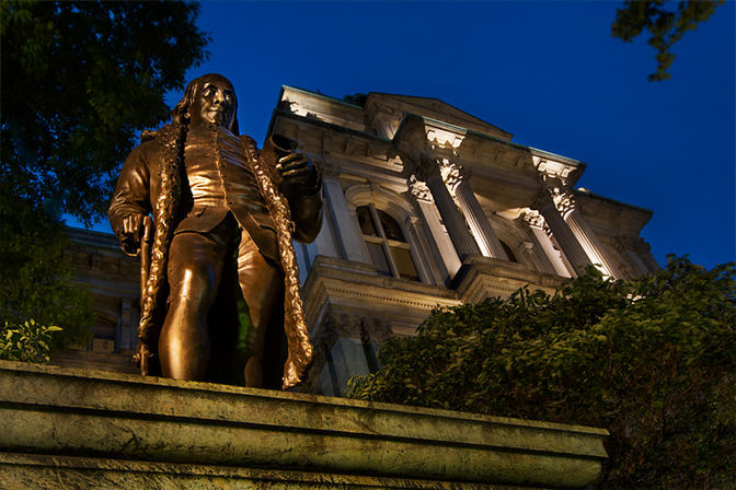 Bronze statue on a stone plinth in front of an illuminated neoclassical building with columns at dusk, framed by trees — historic urban landmark.
