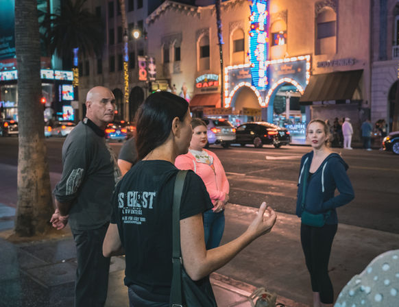 Group of adults talking on a busy, neon-lit theater district sidewalk at night, palm trees, passing cars and an arched marquee glowing in the background.