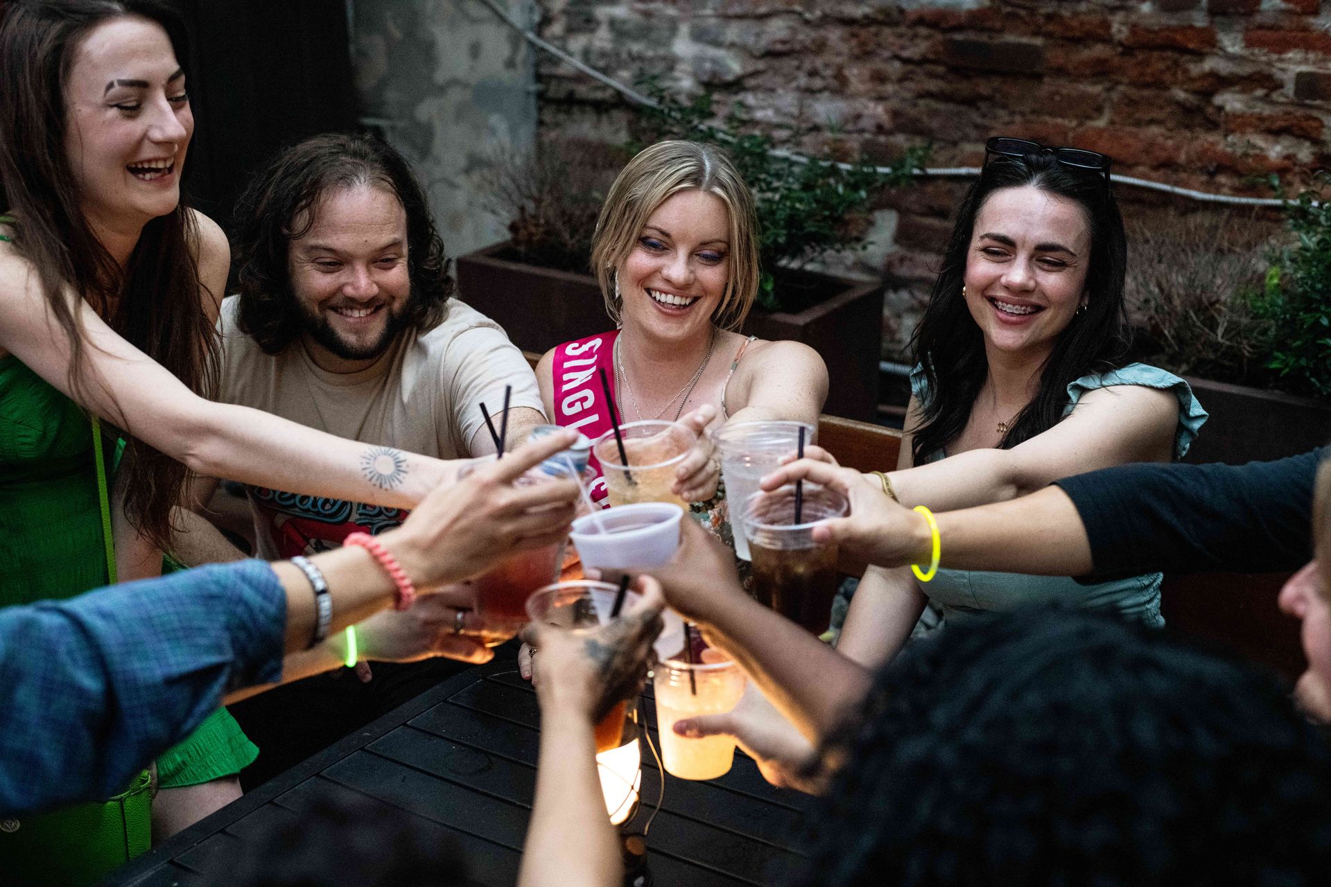 Group of friends toasting with plastic cups on an urban outdoor patio, laughing and smiling around a woman wearing a party sash for a festive bachelorette-style celebration.