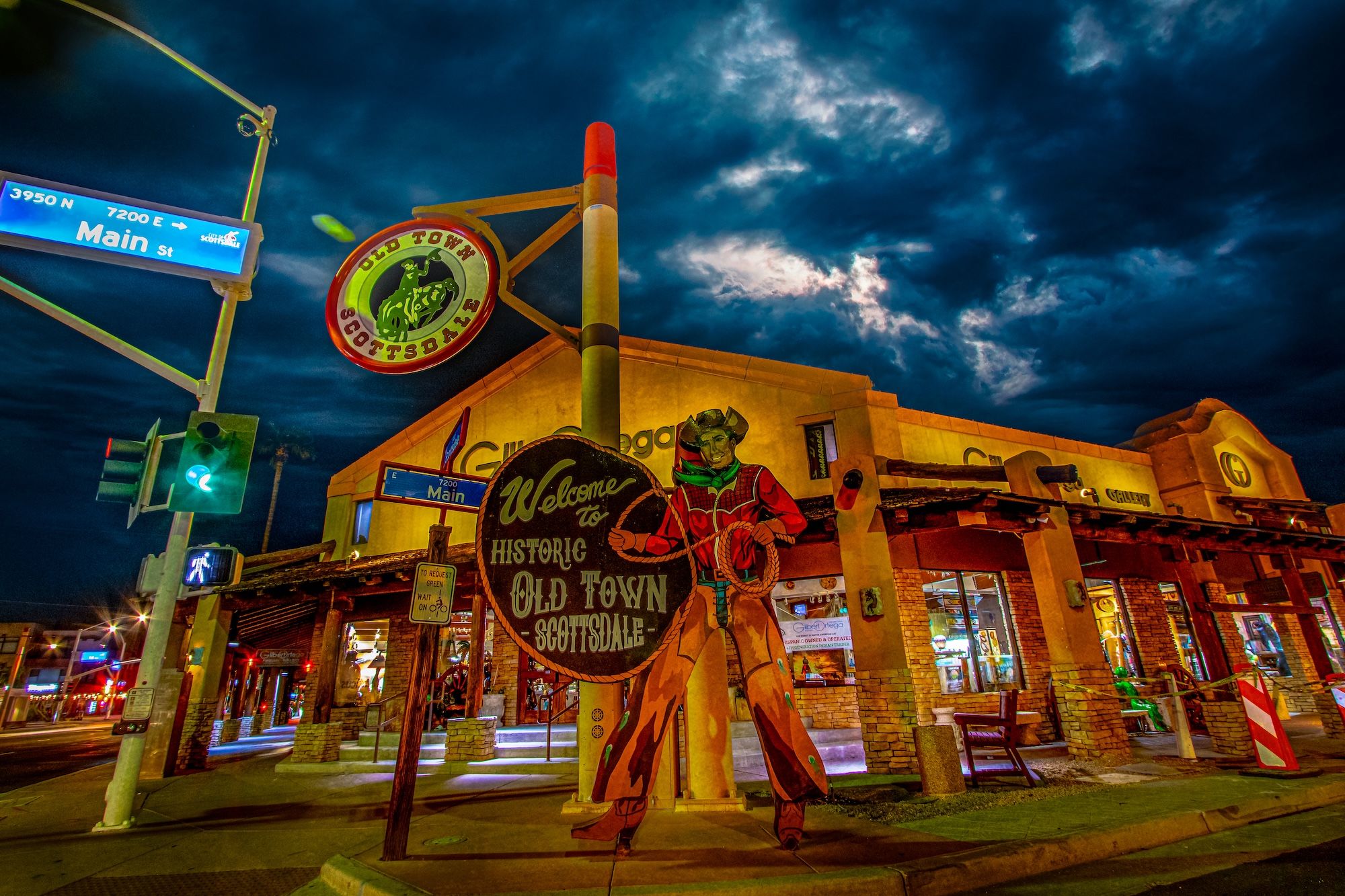 Nighttime street scene in Historic Old Town Scottsdale, Arizona — colorful vintage cowboy welcome sign at the Main Street intersection with illuminated shops and dramatic stormy clouds overhead.