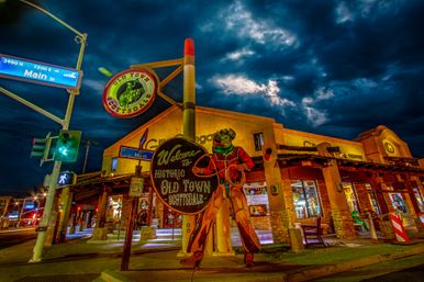 Nighttime street scene in Historic Old Town Scottsdale, Arizona — colorful vintage cowboy welcome sign at the Main Street intersection with illuminated shops and dramatic stormy clouds overhead.