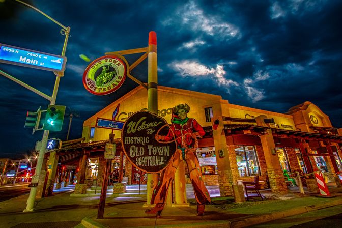Nighttime street scene in Historic Old Town Scottsdale, Arizona — colorful vintage cowboy welcome sign at the Main Street intersection with illuminated shops and dramatic stormy clouds overhead.