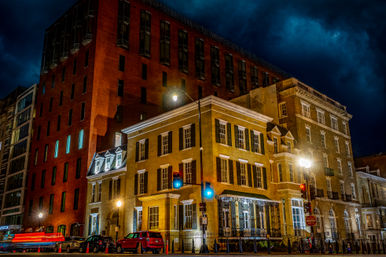 Nighttime downtown street corner with historic brick and brownstone buildings lit by streetlights and traffic signals, parked cars under a dramatic stormy sky