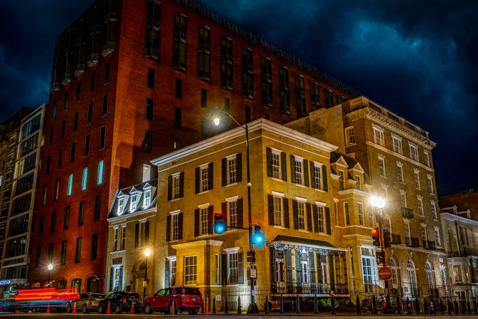 Nighttime downtown street corner with historic brick and brownstone buildings lit by streetlights and traffic signals, parked cars under a dramatic stormy sky