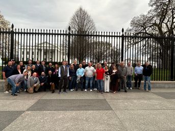 Smiling tour group posing along a tall black iron fence with the White House and early-spring trees behind them in Washington, D.C.
