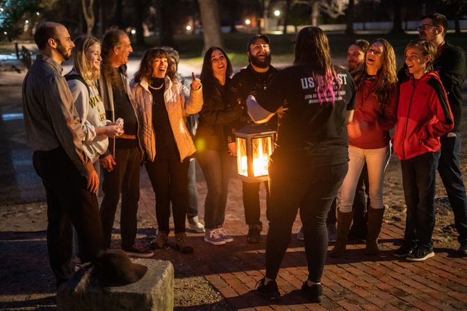 Smiling group gathered around a guide holding a glowing lantern during a lantern-lit nighttime walking tour on a brick path in a city park.