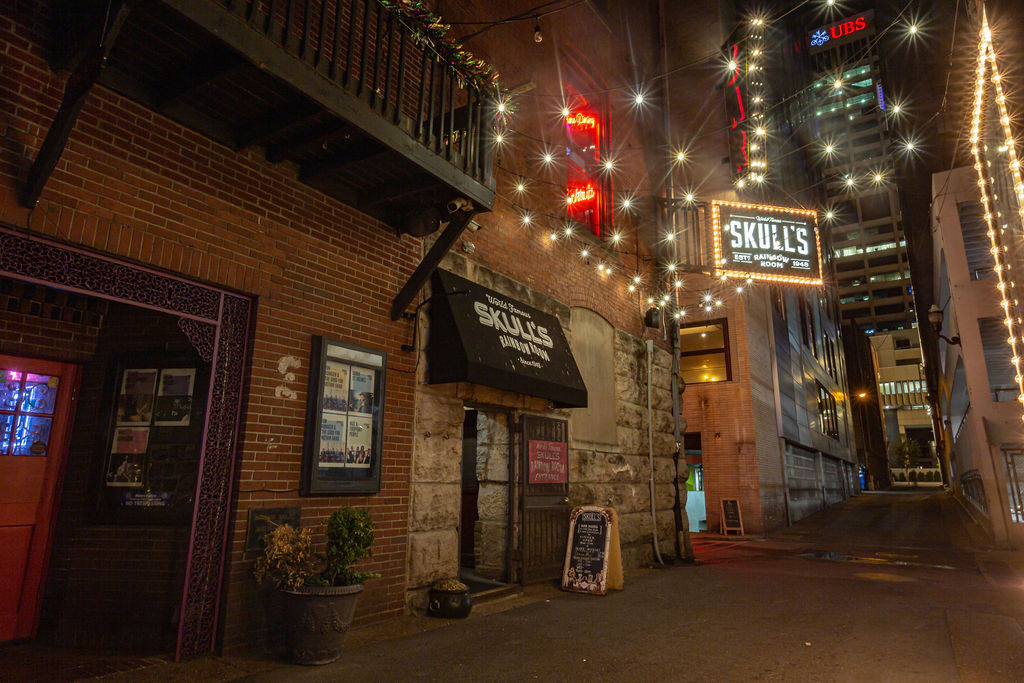Nighttime downtown alley scene with brick storefront and bar entrance under a vintage marquee, twinkling string lights/starburst effects, chalkboard sidewalk sign and high-rise buildings in the background