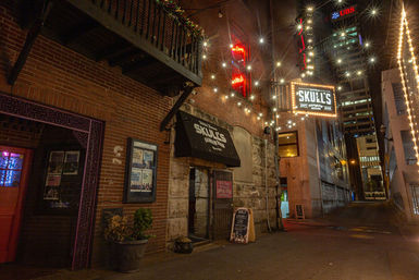 Nighttime downtown alley scene with brick storefront and bar entrance under a vintage marquee, twinkling string lights/starburst effects, chalkboard sidewalk sign and high-rise buildings in the background