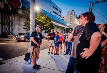Evening city tour on a downtown sidewalk — a group gathered around a guide holding a lantern, with cars, a multi-level parking garage and a chain-link fence in the background.