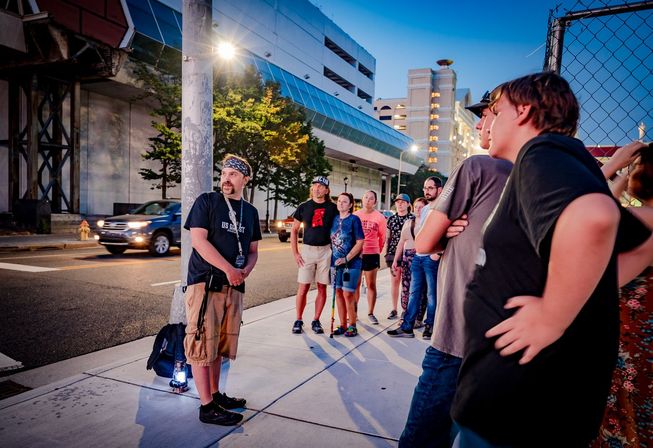Evening city tour on a downtown sidewalk — a group gathered around a guide holding a lantern, with cars, a multi-level parking garage and a chain-link fence in the background.