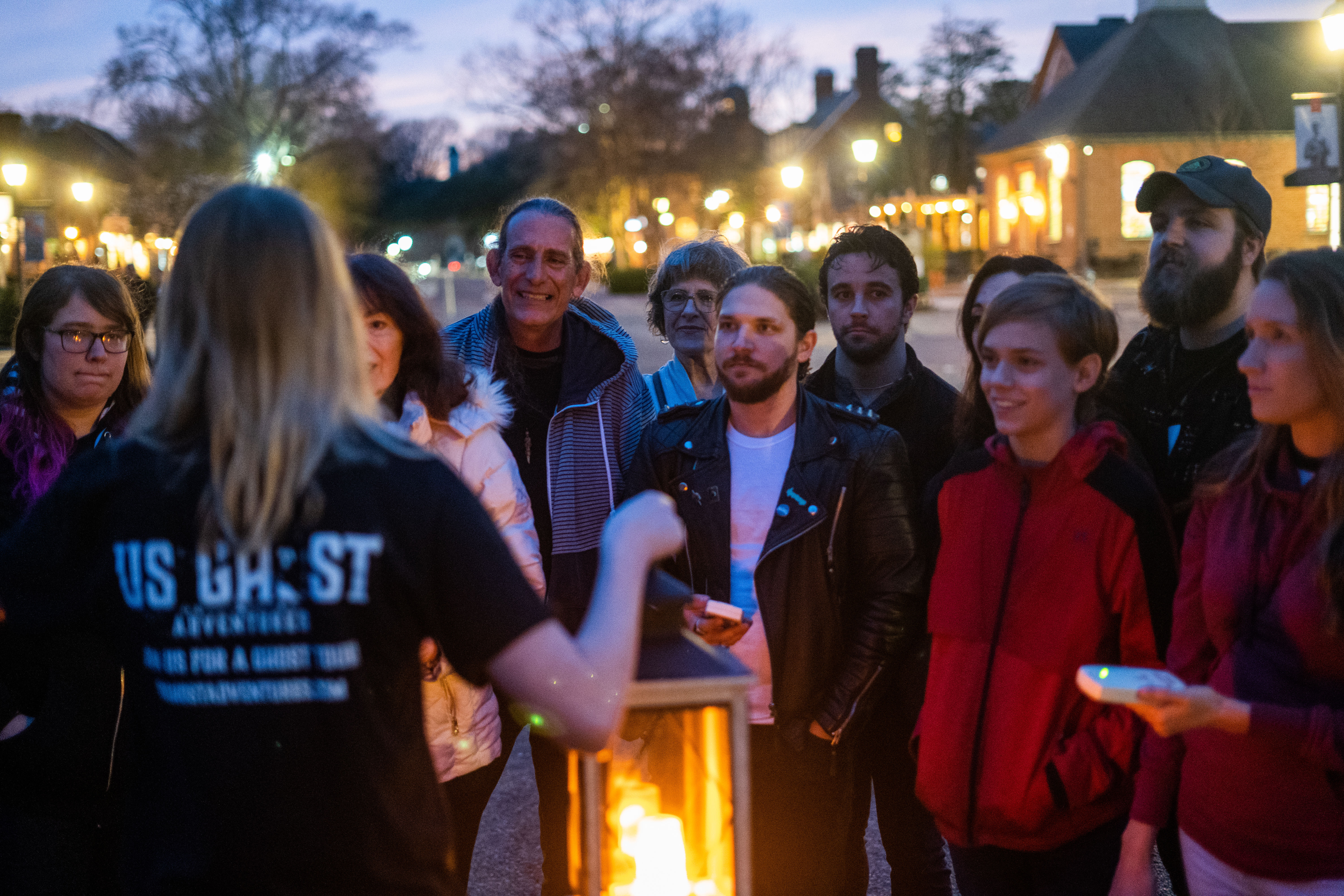 Evening ghost tour group gathered around a glowing lantern on a historic downtown street at twilight, listening to a guide during a small-town walking tour.