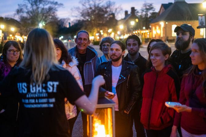Evening ghost tour group gathered around a glowing lantern on a historic downtown street at twilight, listening to a guide during a small-town walking tour.