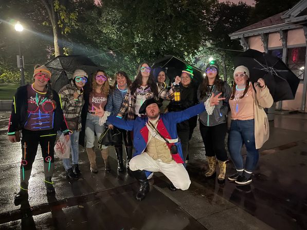Group of friends posing on a wet city sidewalk at night under umbrellas and streetlights, many wearing neon glow glasses and glow-stick accessories, with a man in a blue colonial costume kneeling in front holding a lantern and trees and a park pavilion in the background.