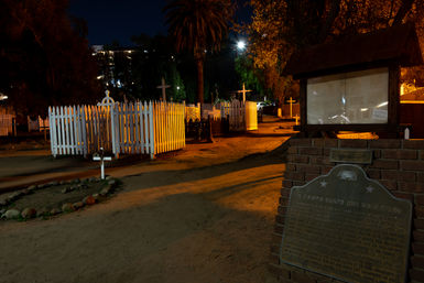 Moody night view of a historic cemetery with white picket-fenced family plots, wooden crosses, a dirt path, palm trees, and a brick memorial plaque bathed in warm streetlight.