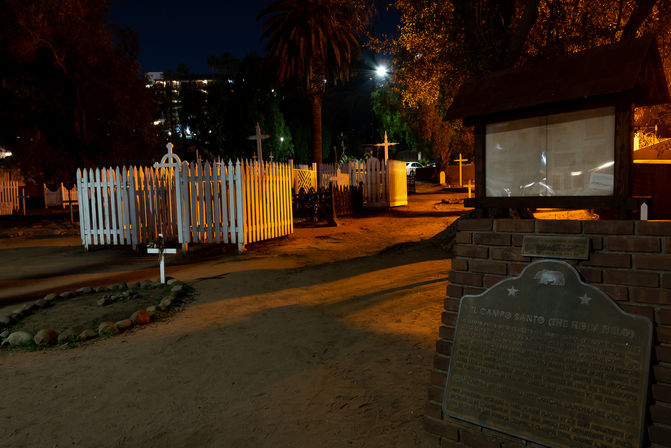 Moody night view of a historic cemetery with white picket-fenced family plots, wooden crosses, a dirt path, palm trees, and a brick memorial plaque bathed in warm streetlight.