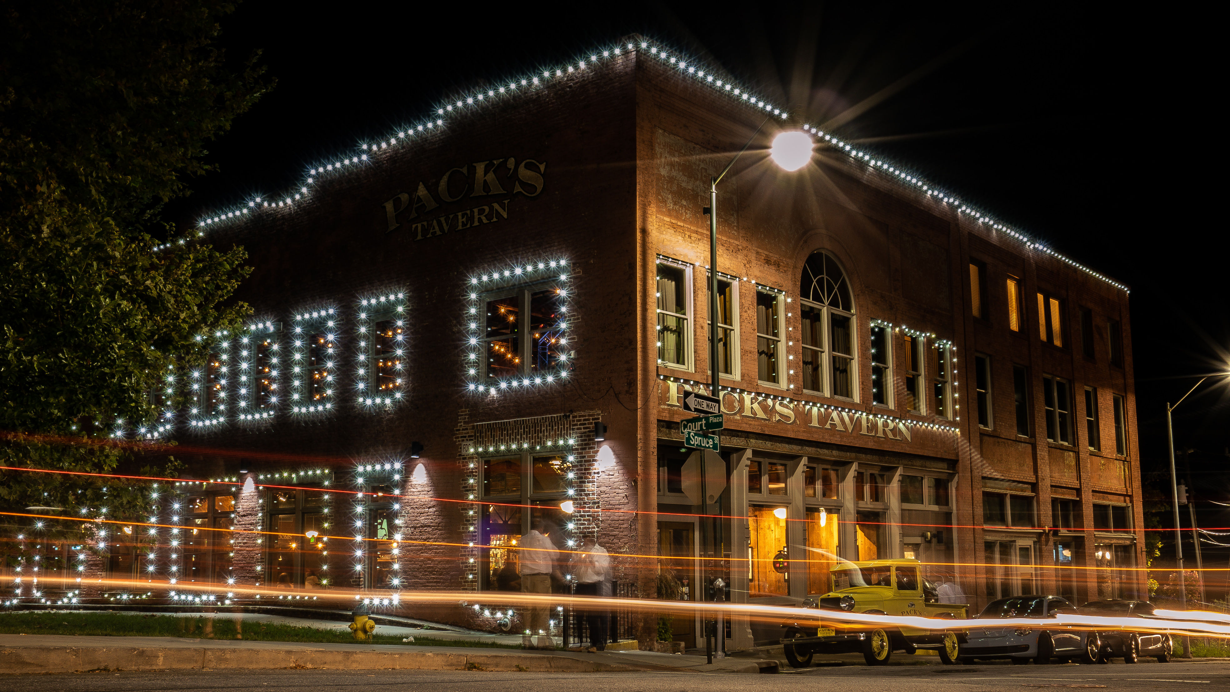 Nighttime downtown brick corner building outlined in white string lights, with streaking car light trails, a vintage pickup parked by the entrance, and a glowing streetlamp.