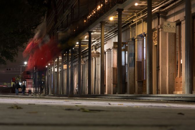 Long-exposure red streetcar streaks past a columned, balcony-lined sidewalk in New Orleans' French Quarter at night, warm shop lights and blurred pedestrians.