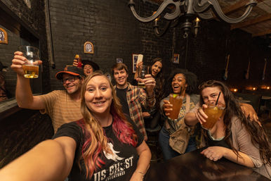 Group selfie of friends toasting with beers and cocktails in a cozy brick‑walled neighborhood bar with exposed beams and a chandelier, lively nightlife vibe