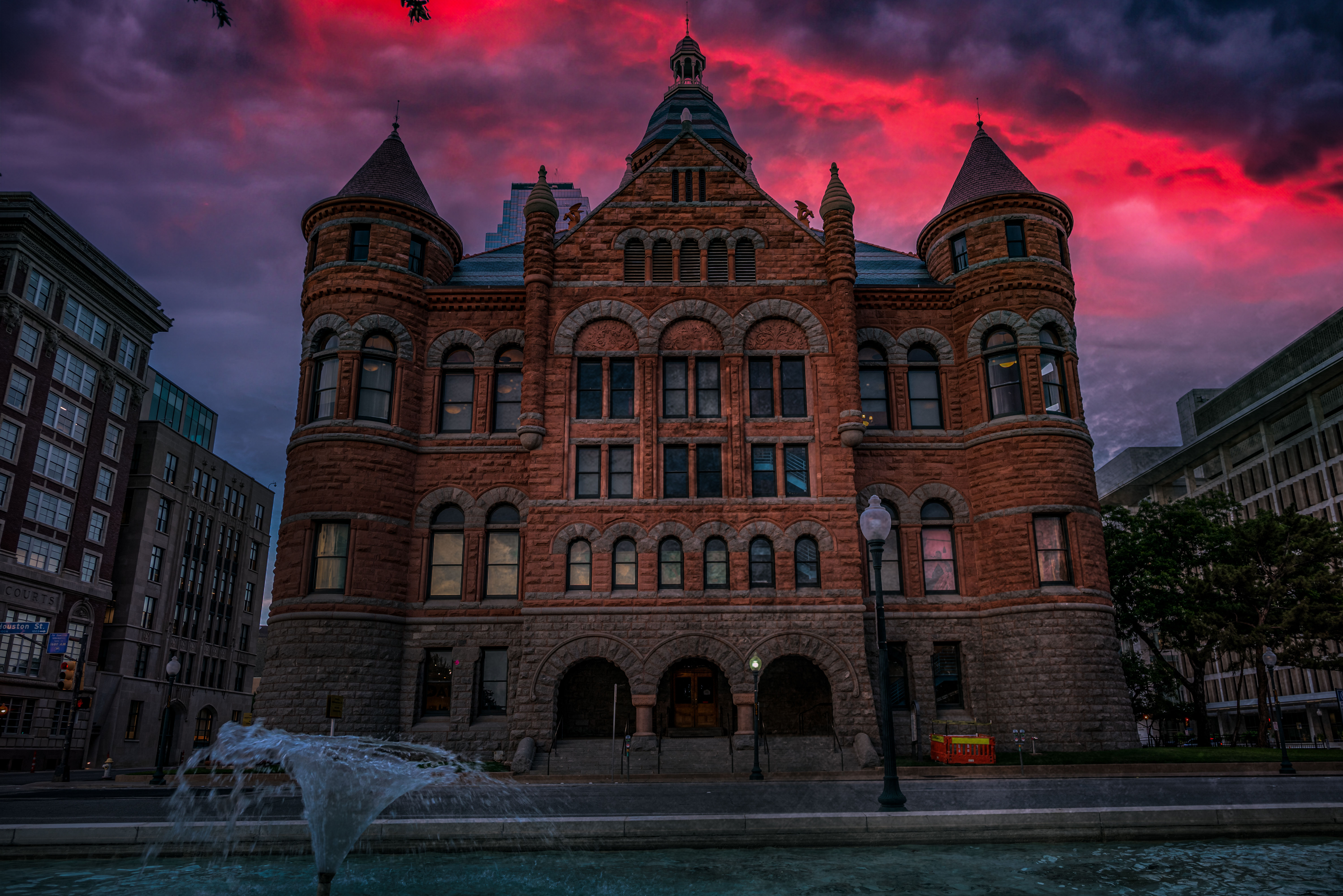 Historic red-brick Romanesque building with round turrets and arched windows beside a fountain in a city plaza under a dramatic magenta sunset sky.