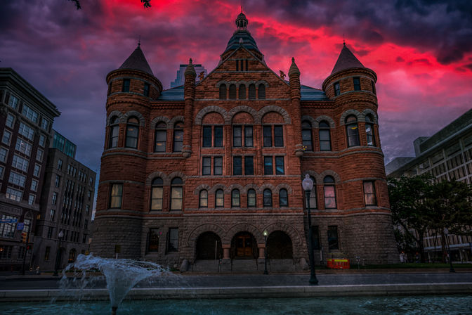 Historic red-brick Romanesque building with round turrets and arched windows beside a fountain in a city plaza under a dramatic magenta sunset sky.