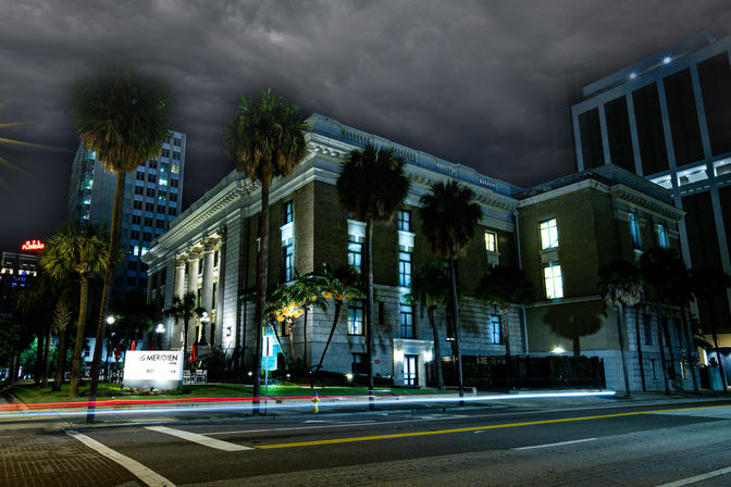 Moody night view of a historic neoclassical downtown building with columns and palm trees, dramatic storm clouds overhead and colorful car light trails along the urban street.