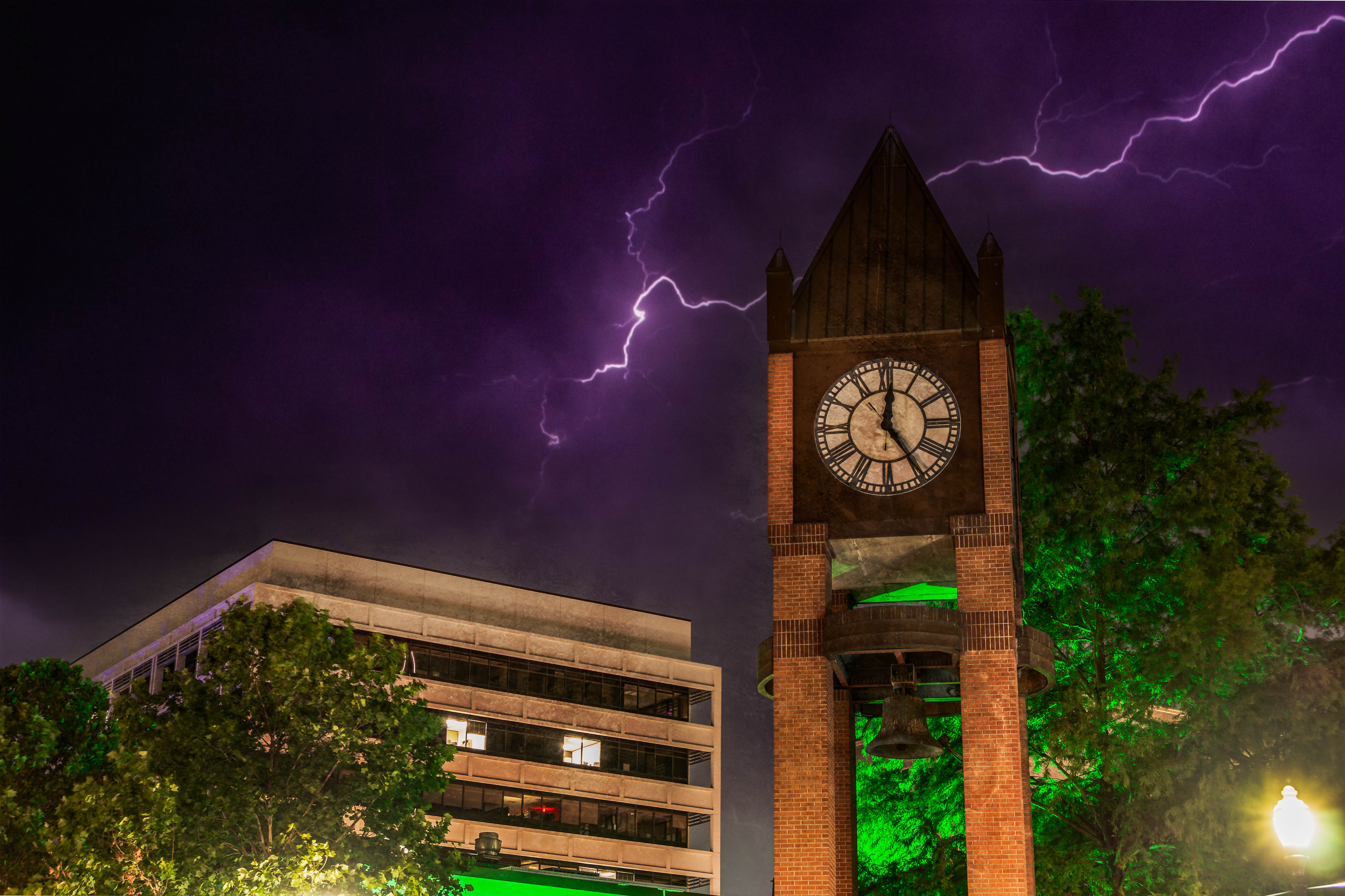 Brick clock and bell tower in a downtown plaza beside an office building, trees lit green under a dramatic purple night sky streaked with lightning.