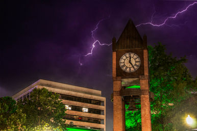 Brick clock and bell tower in a downtown plaza beside an office building, trees lit green under a dramatic purple night sky streaked with lightning.