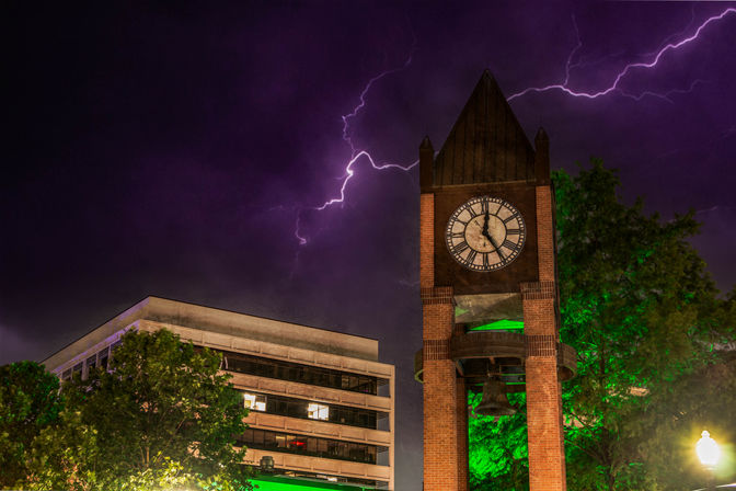 Brick clock and bell tower in a downtown plaza beside an office building, trees lit green under a dramatic purple night sky streaked with lightning.