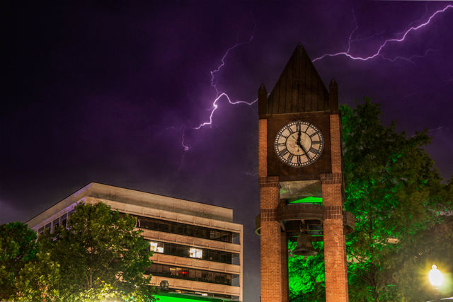 Brick clock and bell tower in a downtown plaza beside an office building, trees lit green under a dramatic purple night sky streaked with lightning.