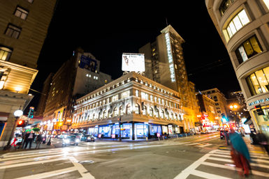 Vibrant nighttime downtown intersection with a brightly lit historic corner building and tall hotel tower, glowing storefronts, moving cars and blurred pedestrians crossing the street.