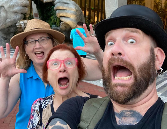 Three adults making exaggerated surprised faces for a selfie — bearded man in a bowler hat, woman with bright pink glasses, and woman in a straw hat — posing in an outdoor plaza in front of a giant stone hands sculpture.