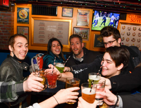 Group of friends laughing and clinking beers and colorful cocktails in a lively sports bar, with a football game on the TV and framed posters on the wall.