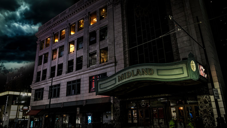 Moody night shot of a historic downtown theater with a curved illuminated marquee, ornate stone facade, and lit upper-floor windows under stormy clouds