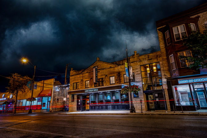 Nighttime downtown scene with a historic stone music-hall façade and neighboring storefronts under a dramatic stormy sky, glowing streetlights, hanging flower baskets, and red long-exposure light trails across an empty road.