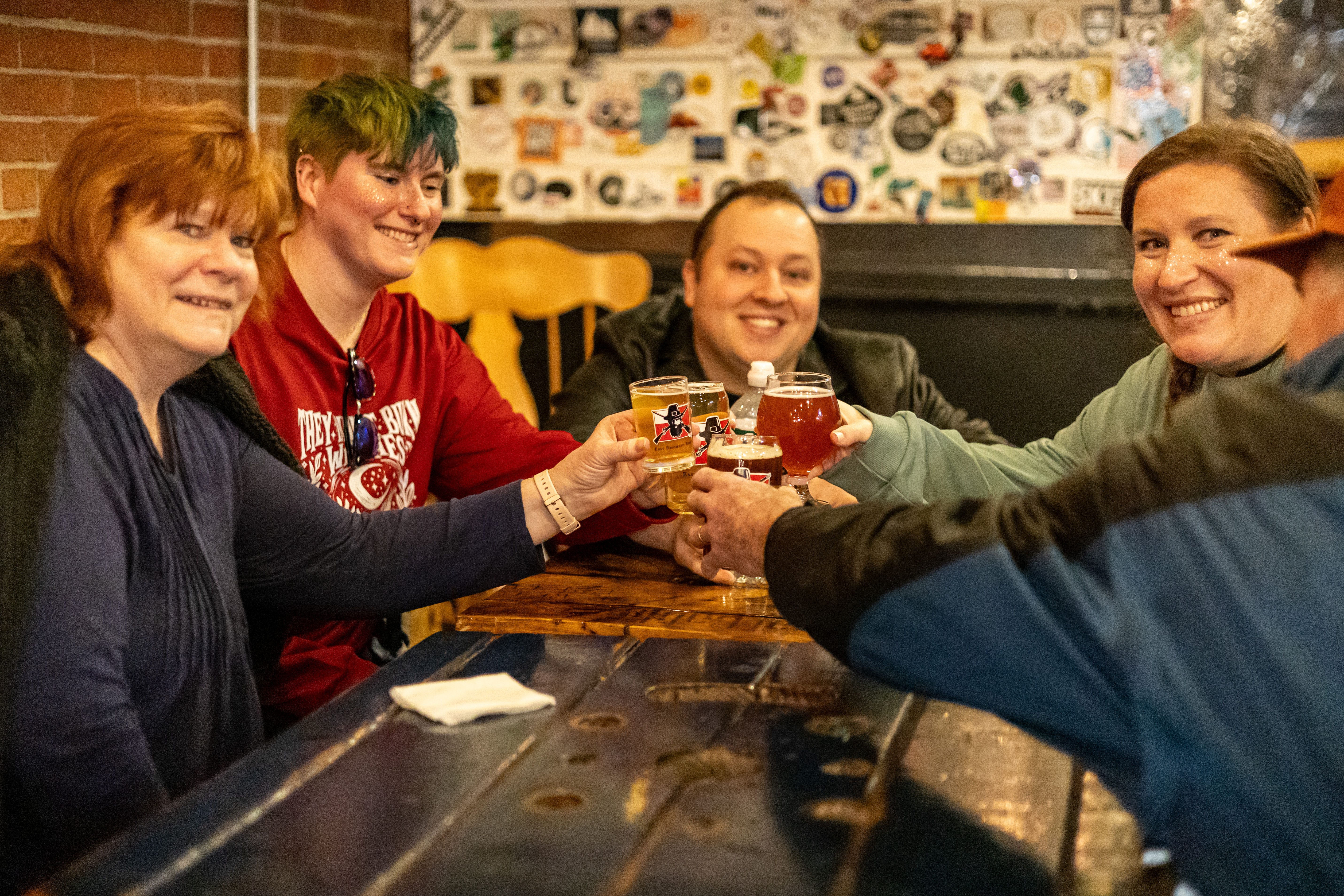 Group of friends toasting craft beers in a cozy brewery taproom at a wooden table with a sticker-covered wall — cheerful beer tasting moment.