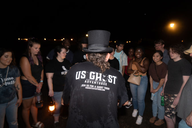 Top-hatted guide leading a lantern-lit nighttime ghost tour on a city street, a diverse group of smiling attendees gathered under streetlights.